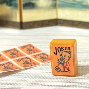 May include: A rectangular mustard-yellow mahjong tile featuring a graphic of a bearded figure in traditional robes with the text JOKER printed above. Beside the tile is a sheet of matching orange stickers with the same JOKER graphic design.