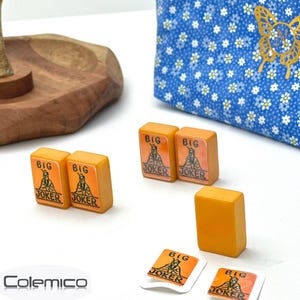 May include: Five orange rectangular mahjong tiles featuring a Big Joker graphic, alongside two small square stickers with the same design. A blue fabric pouch with a white daisy pattern and a gold butterfly emblem sits in the background.