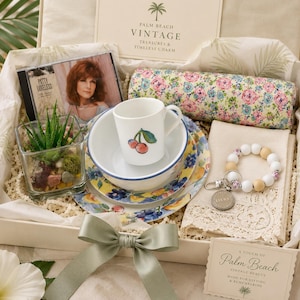 May include: A gift box containing a Patty Loveless CD, a floral print fabric roll, a white mug with a cherry illustration, a bowl and plate with fruit patterns, a succulent in a glass cube, a beaded keychain, and a lace-trimmed cloth napkin.