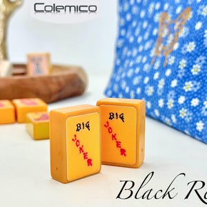 May include: Two orange Mahjong tiles with "BIG JOKER" in black and red text. Other tiles and a blue floral pillow are in the background. The word "Black Red" is written in cursive at the bottom.