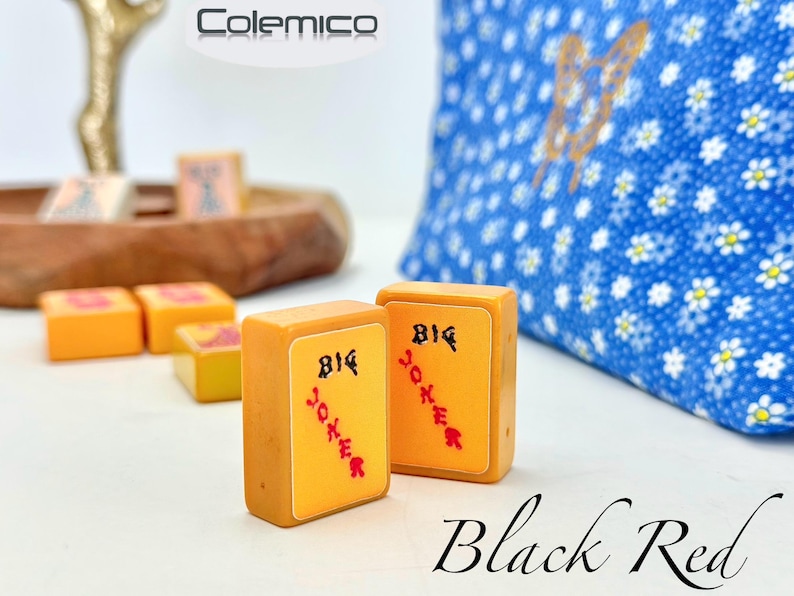 May include: Two orange Mahjong tiles with "BIG JOKER" in black and red text. Other tiles and a blue floral pillow are in the background. The word "Black Red" is written in cursive at the bottom.