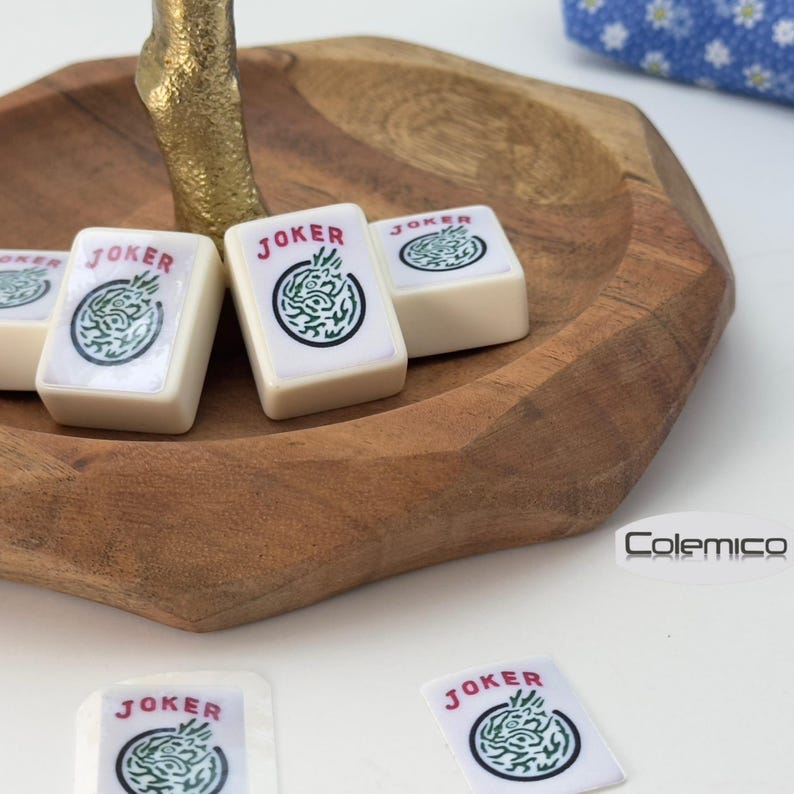 May include: A wooden bowl holds several ivory-colored Mahjong tiles, each marked with the word "JOKER" in red and a green dragon design. The bowl sits on a white surface, with a gold-colored decorative element in the background.