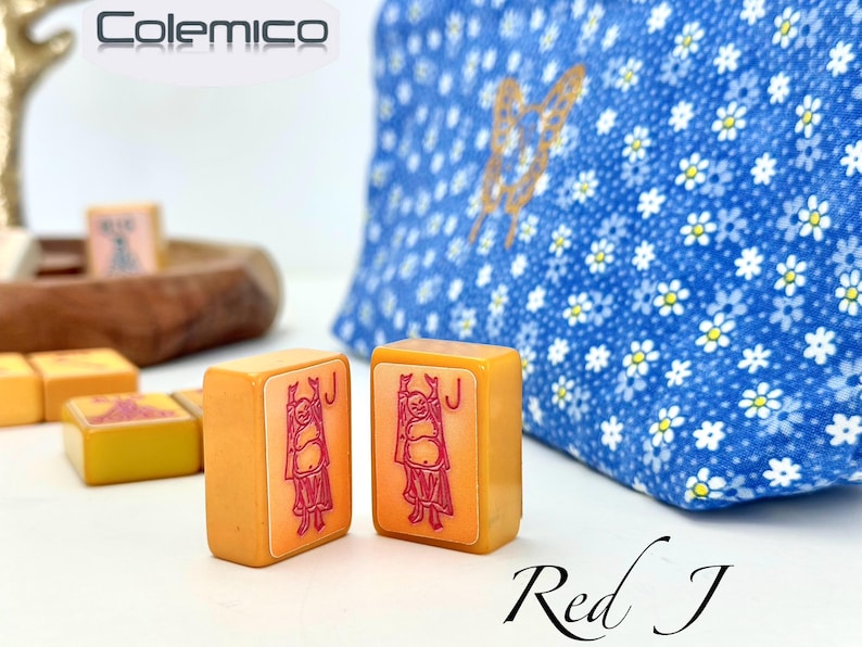 May include: Two orange Mahjong tiles with a red figure and the letter "J" are in focus. A blue floral bag is in the background. The text "Red J" is at the bottom. The brand name "Colemico" is at the top.