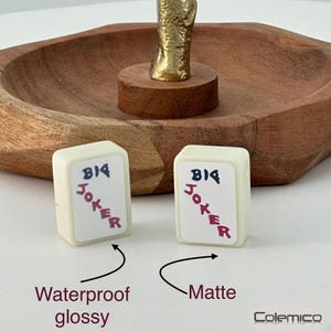 May include: Two small, off-white game tiles with the words "BIG JOKER" in blue and red. The tiles are in front of a wooden tray and a gold-colored decorative element. The text "Waterproof glossy" and "Matte" are also visible.