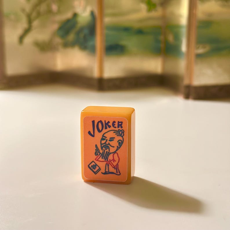 May include: A rectangular mahjong tile with a light orange base colour. The front face features a light peach-coloured inset with a blue and red graphic illustration of a bearded figure and the word JOKER printed in blue capital letters.