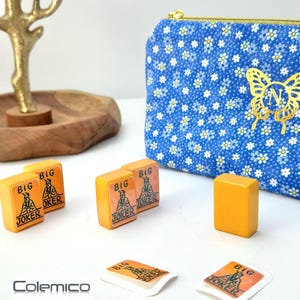 May include: An assortment of orange Mahjong tiles with "BIG JOKER" text and a blue floral print pouch with a gold butterfly design. The tiles are arranged on a white surface, with a wooden tray and gold decorative piece in the background.