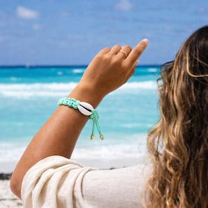 May include: A mint green macrame bracelet with a white seashell centerpiece and gold-colored beads. The bracelet is worn on an arm with a beach and ocean background. The bracelet is a beach-themed accessory.