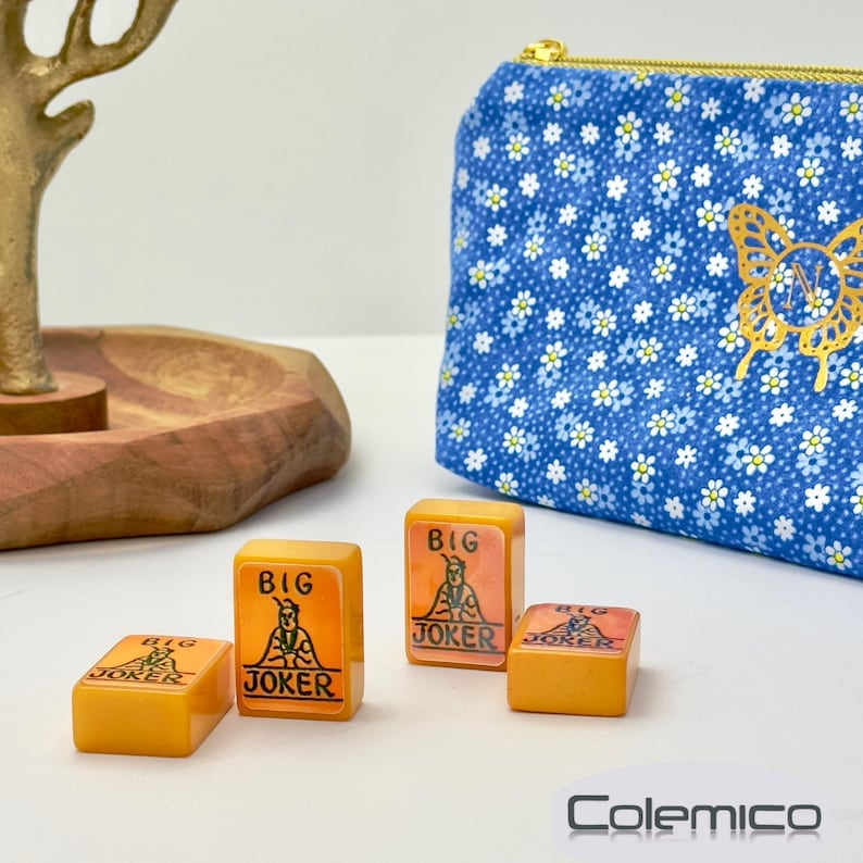 May include: Four orange-yellow Mahjong tiles with "BIG JOKER" text and a graphic of a figure. A blue floral print zippered pouch with a gold butterfly design is in the background. The Colemico logo is at the bottom.