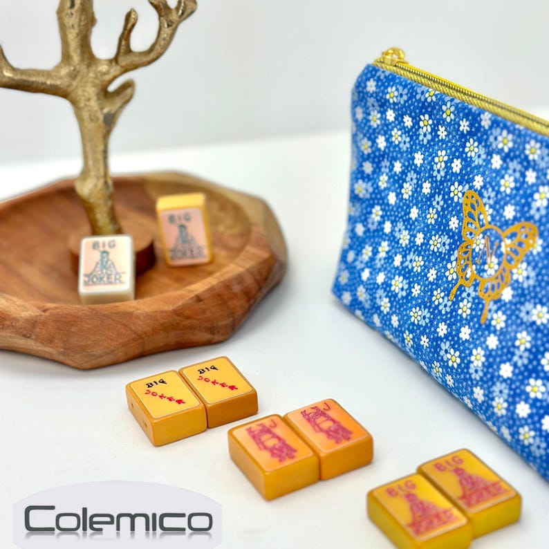 May include: A collection of vintage mahjong tiles and a blue floral print pouch with a gold butterfly design. The tiles are orange and white, some with the word "Joker" and others with Chinese characters. A wooden tray and gold-colored decorative element are also present.
