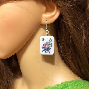May include: A single earring featuring a white square tile with the number 3 and Japanese characters. The tile has a colorful illustration of a person and flowers. The earring is suspended from a silver-colored hook.