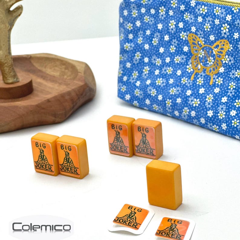 May include: Five orange rectangular mahjong tiles featuring a Big Joker graphic, alongside two small square stickers with the same design. A blue fabric pouch with a white daisy pattern and a gold butterfly emblem sits in the background.