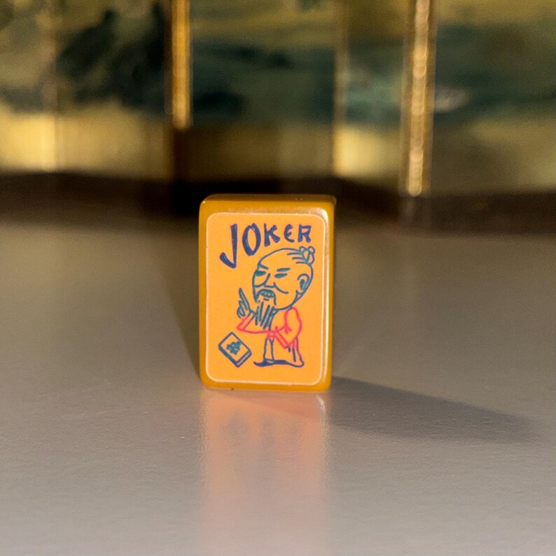 May include: A rectangular yellow mahjong tile featuring a blue graphic of a person with a beard and topknot, the word JOKER in blue text, and a small blue book icon. The tile has a smooth, glossy finish and rounded edges on the front face.