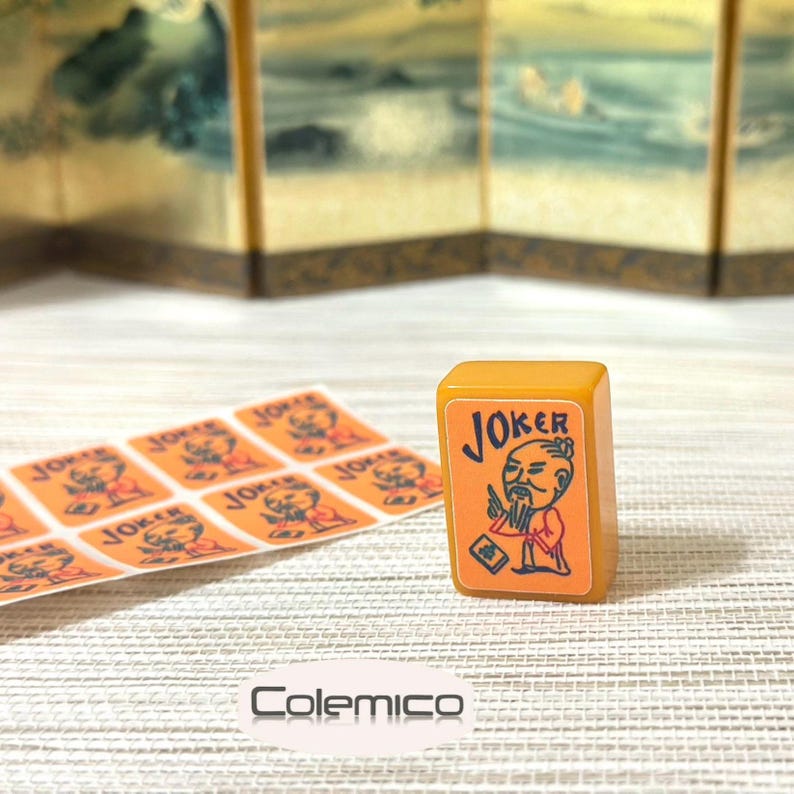 May include: A rectangular mustard-yellow mahjong tile featuring a graphic of a bearded figure in traditional robes with the text JOKER printed above. Beside the tile is a sheet of matching orange stickers with the same JOKER graphic design.