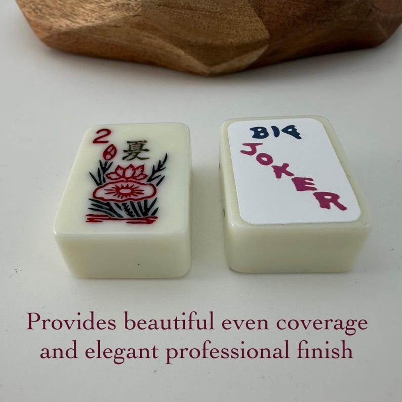 May include: Two ivory-colored Mahjong tiles. One tile features a red lotus flower design with the number 2 and Chinese characters. The other tile displays the word "BIG JOKER" in blue and pink. The tiles are on a white surface.