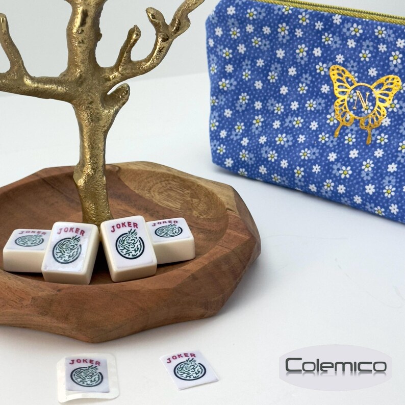May include: A wooden bowl holds several white Mahjong tiles with "JOKER" text and a green design. A blue floral patterned pouch with a gold butterfly design and zipper is in the background. A gold-colored decorative tree is also present.
