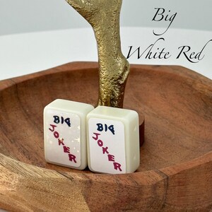May include: Two white game tiles with "BIG JOKER" in red and black lettering. The tiles are in a wooden bowl with a gold-colored decorative element. The text "Big White Red" is written in cursive.