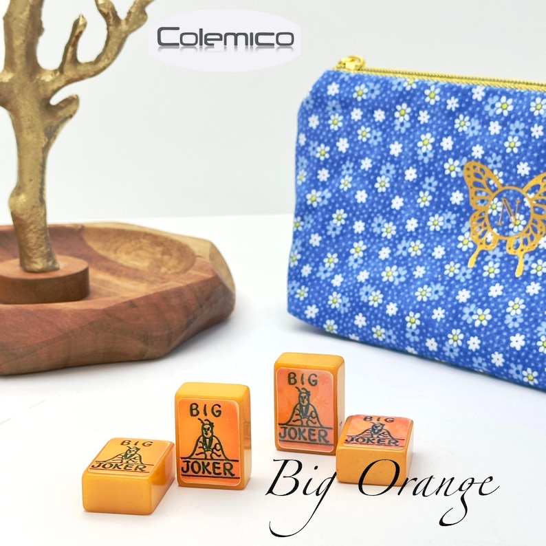 May include: Four orange Mahjong tiles with "BIG JOKER" text and a graphic of a joker. A blue floral print pouch with a gold butterfly design is in the background. The words "Big Orange" are written in cursive.