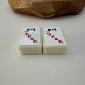 May include: Two rectangular, off-white playing card-shaped objects with the words "BIG JOKER" printed on them in blue and red. The objects are set against a white background, with a wooden bowl in the background.