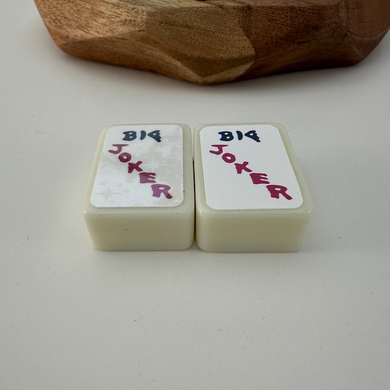May include: Two rectangular, off-white playing card-shaped objects with the words "BIG JOKER" printed on them in blue and red. The objects are set against a white background, with a wooden bowl in the background.