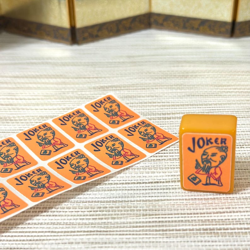 May include: A rectangular mahjong tile with an orange face featuring a black and red illustration of a bearded figure and the word JOKER, accompanied by a sheet of matching orange stickers with the same graphic design and text printed on them.