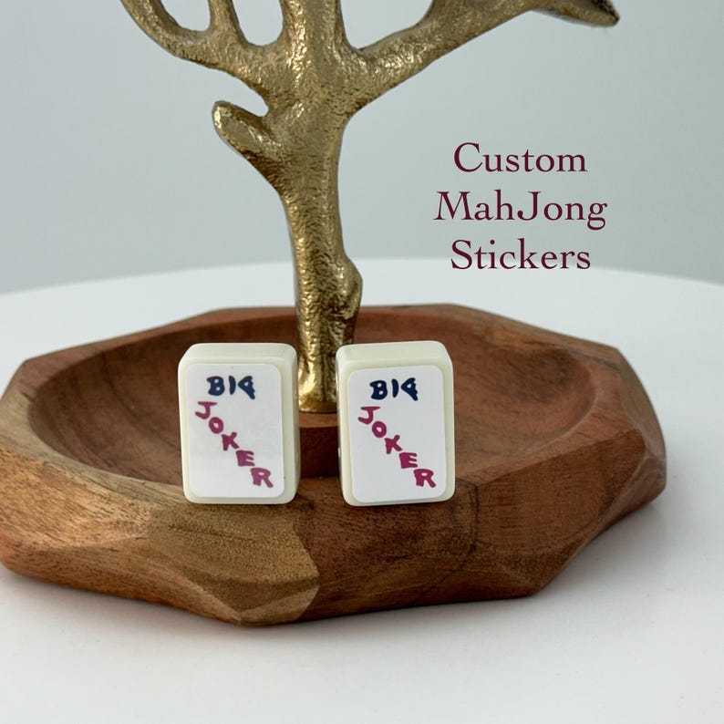 May include: Two off-white Mahjong tiles with "BIG JOKER" text in blue and red, displayed on a wooden tray. The tiles are part of a set of custom Mahjong stickers. A gold-colored decorative element is behind the tray.