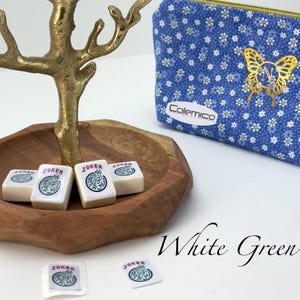 May include: A wooden bowl holds white Mahjong tiles with green and black "Joker" symbols. A gold-colored decorative tree stands above the bowl. A blue floral print pouch with a gold butterfly design and the brand name "Colemico" is also present.