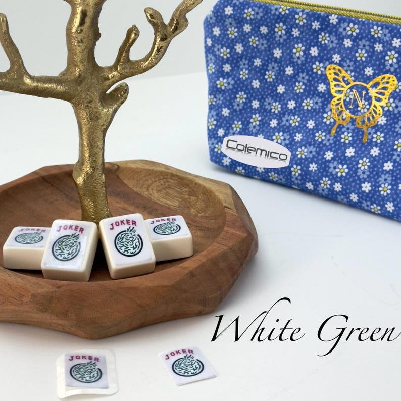 May include: A wooden bowl holds white Mahjong tiles with green and black "Joker" symbols. A gold-colored decorative tree stands above the bowl. A blue floral print pouch with a gold butterfly design and the brand name "Colemico" is also present.