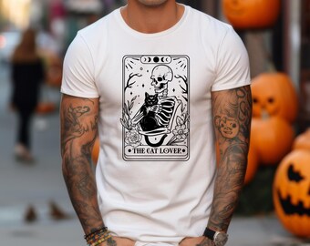 Puede incluir: Camiseta blanca con un gráfico en blanco y negro de un esqueleto sosteniendo un gato negro, con el texto "THE CAT LOVER". El diseño incluye elementos florales y una ilustración de fases lunares. Una prenda casual.