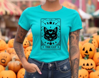 Puede incluir: Una camiseta turquesa con un diseño de carta de tarot de gato negro. El diseño incluye la cara de un gato, una luna creciente, estrellas y el texto "THE CAT". La camiseta está hecha de un material suave y es adecuada para uso casual.