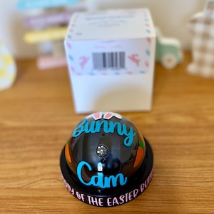 May include: A black bell with the words "Bunny Cam" in blue script and orange carrot graphics. The base reads "Day of the Easter Bunny" in pink. A white box is in the background.