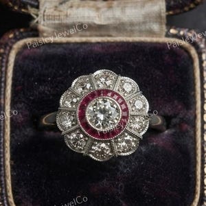 May include: A silver-toned ring featuring a central round diamond surrounded by a halo of small red stones and an outer scalloped border of round diamonds. The ring is set in a metal band and presented inside a dark purple velvet-lined jewelry box.