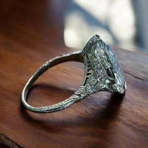 Könnte beinhalten: Ein antiker Silberring mit einem großen, rechteckigen, klaren Edelstein. Der Ring weist aufwendige Filigranarbeiten und kleine Akzentsteine am Band und der Fassung auf. Der Ring wird auf einer dunklen Holzoberfläche präsentiert.