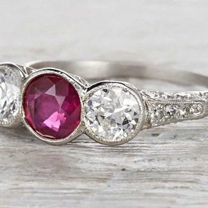 May include: A vintage-style engagement ring featuring a large, deep red ruby flanked by two smaller diamonds. The band is set with smaller diamonds and has an intricate, engraved design.
