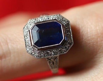 May include: A silver ring with a large, dark blue gemstone surrounded by smaller, clear gemstones.