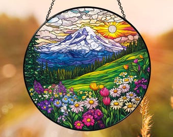 Könnte beinhalten: Ein runder Buntglas-Sonnenfänger mit einem schneebedeckten Berggipfel unter einem Sonnenaufgangshimmel. Der Vordergrund zeigt eine lebendige Wiese voller bunter Wildblumen, darunter Gänseblümchen, Tulpen und violette Blüten, eingefasst von einem schwarzen Metallrahmen.