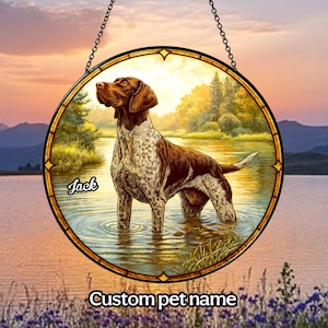 May include: A stained glass suncatcher featuring a brown and white German Shorthaired Pointer dog standing in a lake. The dog is looking to the left and the suncatcher has a gold frame. The text "Jack" is written on the suncatcher.