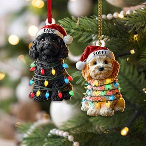 May include: Two dog-shaped Christmas ornaments. One is black, the other is tan. Both wear Santa hats and are wrapped in colorful Christmas lights. The black dog ornament is labeled "Daisy", and the tan dog ornament is labeled "Poppy".