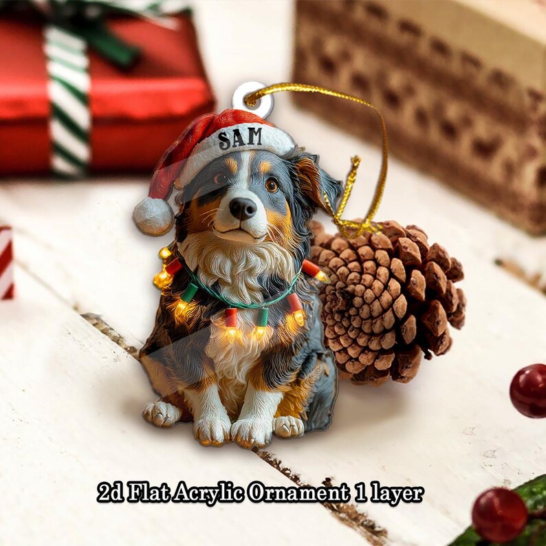 May include: A 2D flat acrylic ornament featuring a dog wearing a Santa hat with the name "SAM" and a string of lights. The dog is black, brown, and white. The ornament is hanging with a gold string next to a pinecone.