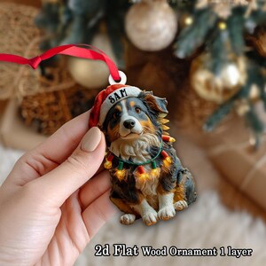 May include: A Christmas ornament shaped like a dog wearing a Santa hat with the name "SAM" on it. The dog has a festive string of lights around its neck. The ornament is made of wood and has a red ribbon for hanging.