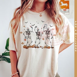 May include: A white t-shirt with a graphic of three skeletons dressed as nurses. The skeletons are holding medical supplies and are surrounded by fall leaves and pumpkins.