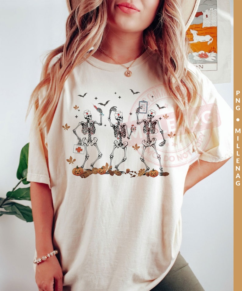 May include: A white t-shirt with a graphic of three skeletons dressed as nurses. The skeletons are holding medical supplies and are surrounded by fall leaves and pumpkins.