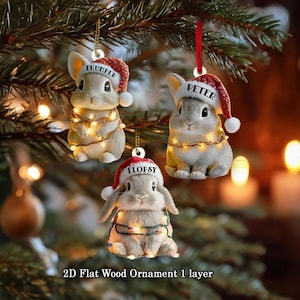 May include: Three Christmas ornaments shaped like bunnies, each wearing a Santa hat with a name on it: Thumper, Peter, and Flopsy. The bunnies are wrapped in string lights and are hanging from a Christmas tree. The text "2D Flat Wood Ornament 1 layer" is at the bottom.