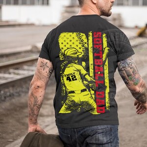 May include: Black t-shirt with a yellow and red graphic design. The graphic features a softball player with the text "Softball Dad" and "Emily 42".