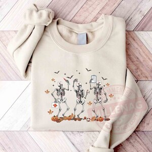 May include: Three skeleton nurses wearing scrubs and hats are holding medical supplies. They are surrounded by fall leaves, pumpkins, and bats. The skeletons are smiling and appear to be happy.