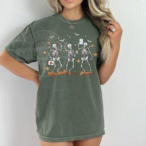 May include: A green t-shirt with a graphic of three skeletons dressed as nurses. The skeletons are holding medical supplies and are surrounded by fall leaves and pumpkins. The text "Nurse Life" is printed on the shirt.