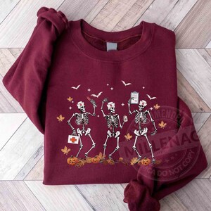 May include: A maroon sweatshirt with a graphic of three skeletons dressed as nurses. The skeletons are holding medical supplies and a cup of coffee. The graphic is surrounded by fall leaves and pumpkins.
