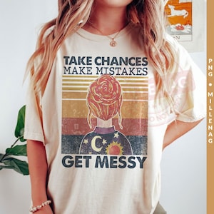 Könnte beinhalten: Ein beiges T-Shirt mit einer Grafik im Retro-Stil, die eine Frau mit einem Dutt, einen zunehmenden Mond und eine Sonne zeigt. Der Text auf dem Shirt lautet "Take Chances Make Mistakes Get Messy".