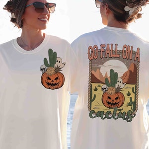 May include: White t-shirt with a graphic of a jack-o'-lantern in a cactus pot with a ghost and skulls. The back of the shirt has the text "Go Fall On A Cactus" with a graphic of a jack-o'-lantern in a cactus pot with a ghost and skulls.