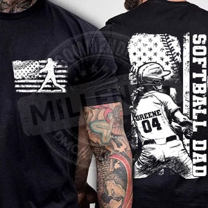 Op de afbeelding: Zwart T-shirt met een witte afbeelding van een softbalspeler voor een Amerikaanse vlag met de tekst "Softball Dad".