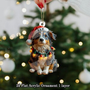 May include: A 2D flat acrylic Christmas ornament featuring a dog wearing a Santa hat with the name "SAM" and a string of lights. The dog is black, brown, and white. The ornament is hanging in front of a blurred Christmas tree.